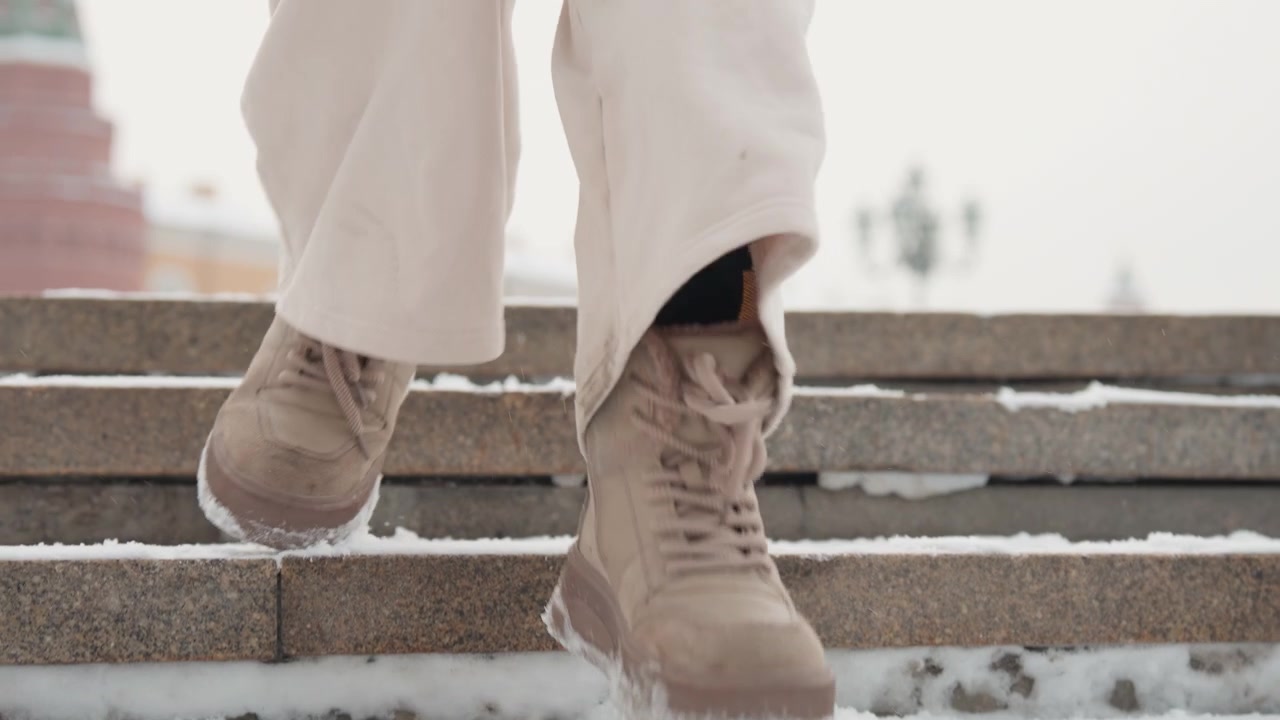 Low-angle view of lower legs and lace-up boots descending outdoor concrete steps with scattered snow and slush on edges, showing cold winter movement, texture, and urban atmosphere