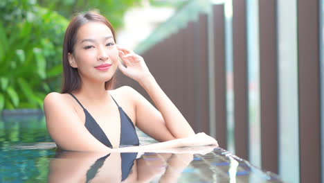 a pretty young fit woman is leaning on the edge of a resort's swimming pool enjoying the water, she is looking at the camera lens with attraction and desire smi