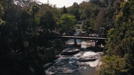 huge large waterfalls under the bridge where a van drives over in mae klang waterfalls among the green trees in thailand on a summer afternoon