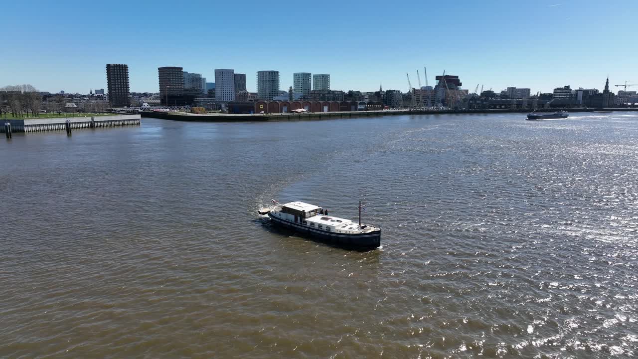 Zoom out elevating aerial shot of a boat on Antwerp river with modern city skyline and harbor cranes in the background under blue sky