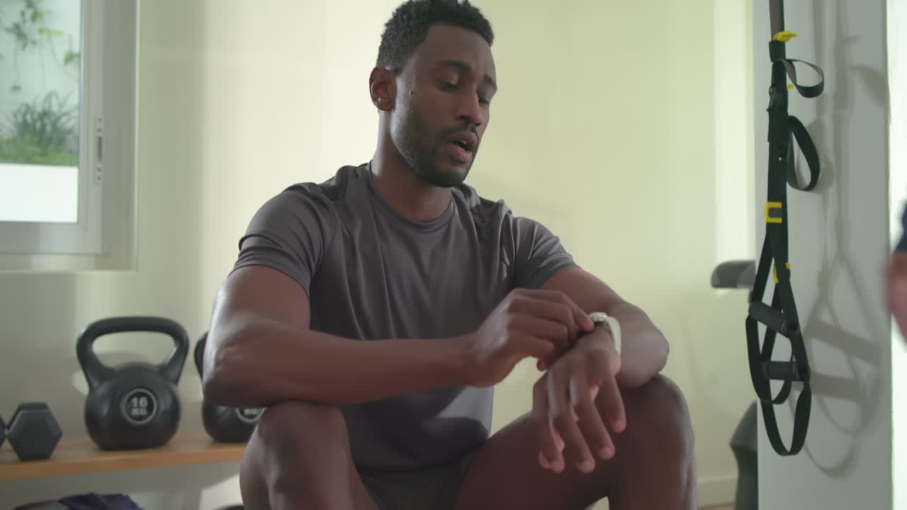 African American man rowing checking watch, sipping water while friend coaching at gym, copy space