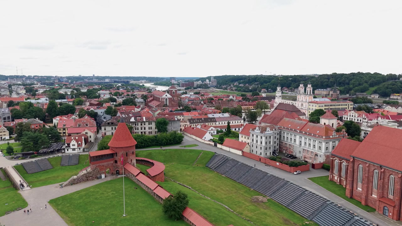 Splendide riprese aeree rivelano l'architettura con tetti rossi della Città Vecchia di Kaunas e l'iconico Castello di Kaunas, circondati da una vegetazione lussureggiante, sotto la luce del giorno con un'illuminazione morbida e uniforme.