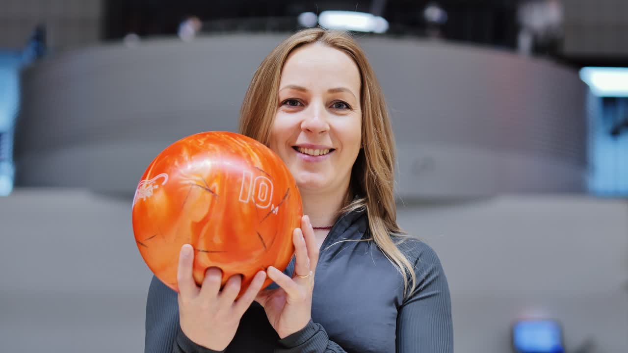 Focused woman smiles holding orange bowling ball in slow motion portrait moment
