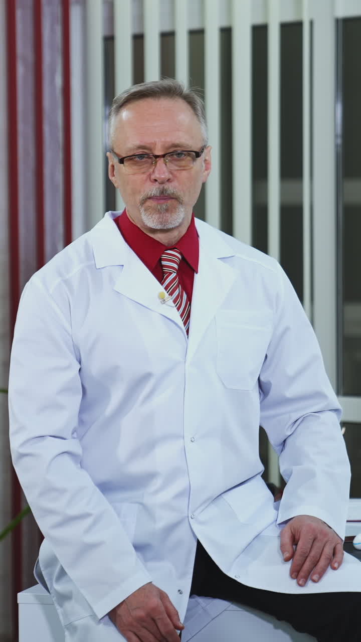 Physician recording webinar training. Professional male doctor sitting on desk and talking in healthcare center. Doctor in glasses and white medical uniform looking at camera. Vertical video