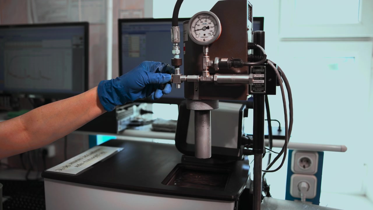 Close view shows gloved technician hand adjusting small metal valve on pressure testing rig with gauge, tubing, stand, and fittings inside clean industrial workspace with cool light