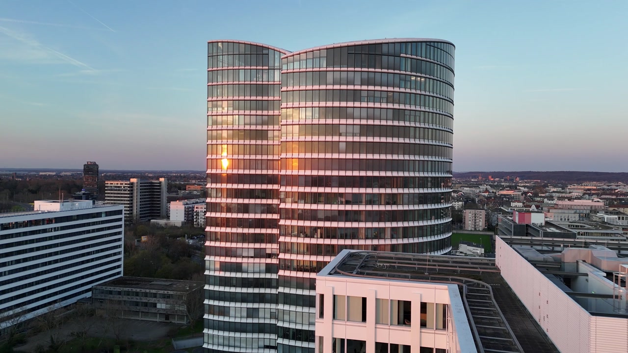 Drone flies across the Rhine toward the double oval Sky Office towers glowing in orange sunset.