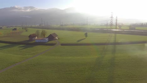 Panorámica-De-Un-Campo-Verde-A-Una-Casa-Con-Una-Granja,-La-Chimenea-Está-Humeando