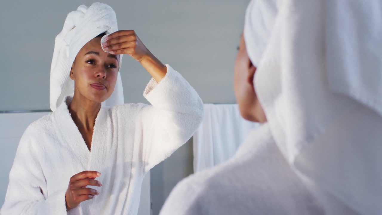 African american woman in bathrobe cleaning her face with cotton pad looking in the mirror at bathro
