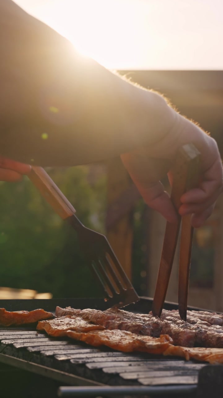 Vertical video shows close-up, sunlit hands grilling meat outdoors, capturing smoky barbecue vibes, golden hour light, and mobile-friendly composition.