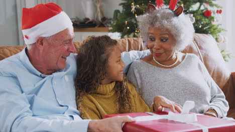 Excited-Granddaughter-Sitting-On-Sofa-With-Grandparents-Opening-Presents-On-Christmas-Day