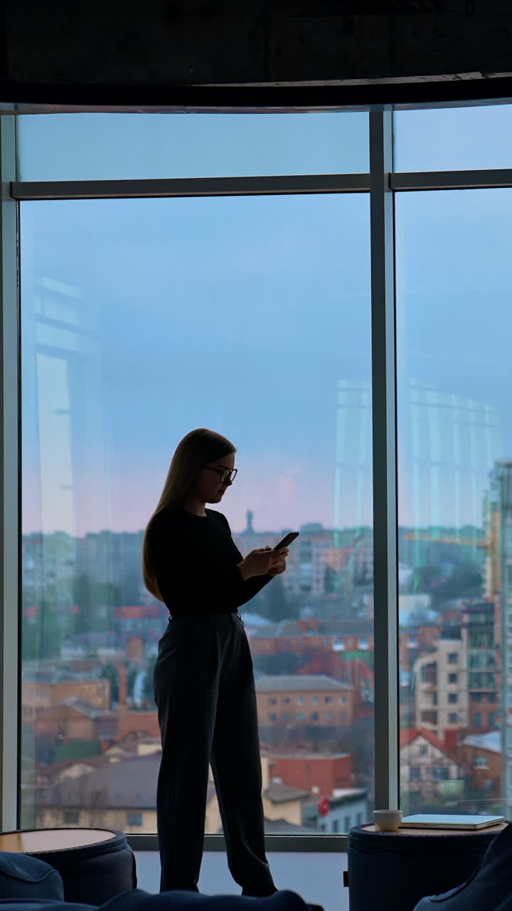 Full-length portrait on a business woman in modern office. Panoramic view on large office windows with cityscape indoors. Beautiful female business worker with a phone. Vertical video