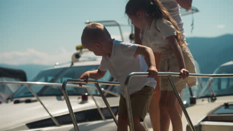 toddler boy looks down from yacht drawbridge landing with mother and sister in city port. junior brother scared to go down vessel ladder with family