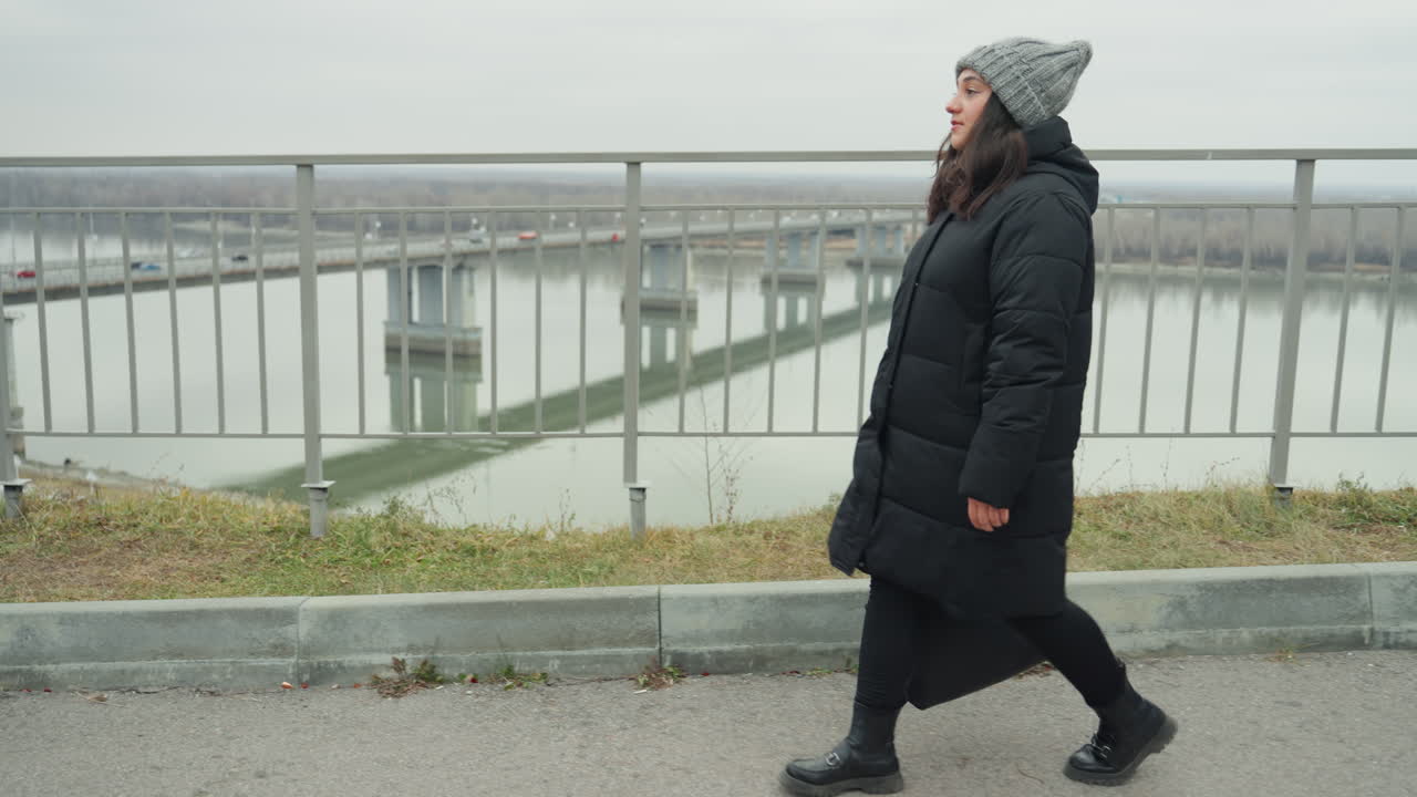 Vrouw wandelt langs de reling van een rivier, gekleed in een gewatteerde jas en gebreide muts. Onder een bewolkte lucht, met een brug en water op de achtergrond, zet ze langzame, gestage stappen. Een peinzende stemming, koude lucht en gedempte tinten kenmerken de stedelijke waterkant.