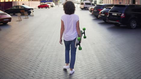 vista posterior de una mujer caminando por la calle de la ciudad vieja sosteniendo su longboard por la mañana, aparcando en la ciudad. hermosa vieja