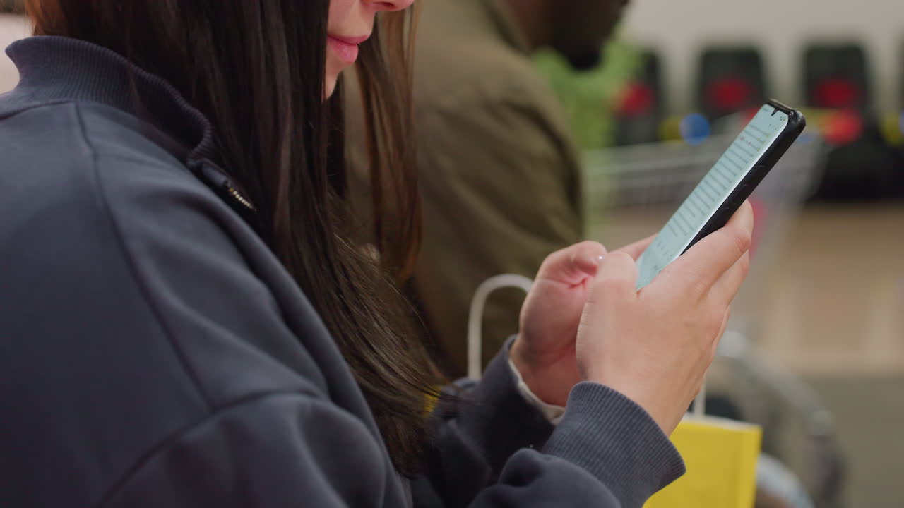 Close up of woman typing on smartphone while sitting indoors beside another person, showing digital interaction and modern lifestyle with focus on screen and hands, slight blur in background