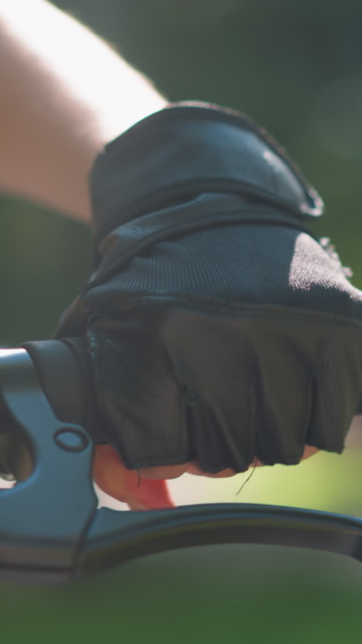 장갑을 입은 손으로 개별적으로 잡는 자전거 손잡이의 클로즈업, 가속화 및 브레이크를 잡고, 빛이 손에서 반사되며, 배경에는 부드럽게 흐릿한 초록색이 있습니다.