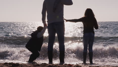 Vista-Trasera-Del-Padre-Con-Hijos-Saltando-Sobre-Las-Olas-Mirando-Al-Mar-Siluetas-Contra-El-Sol.