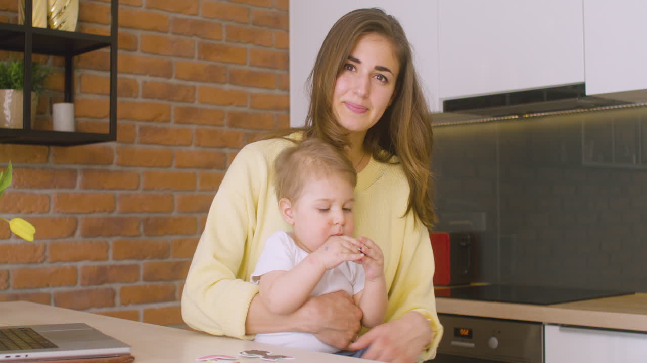 Woman Looking At Camera While Is Sitting In The Kitchen And Holding Her Baby On Her Lap 1