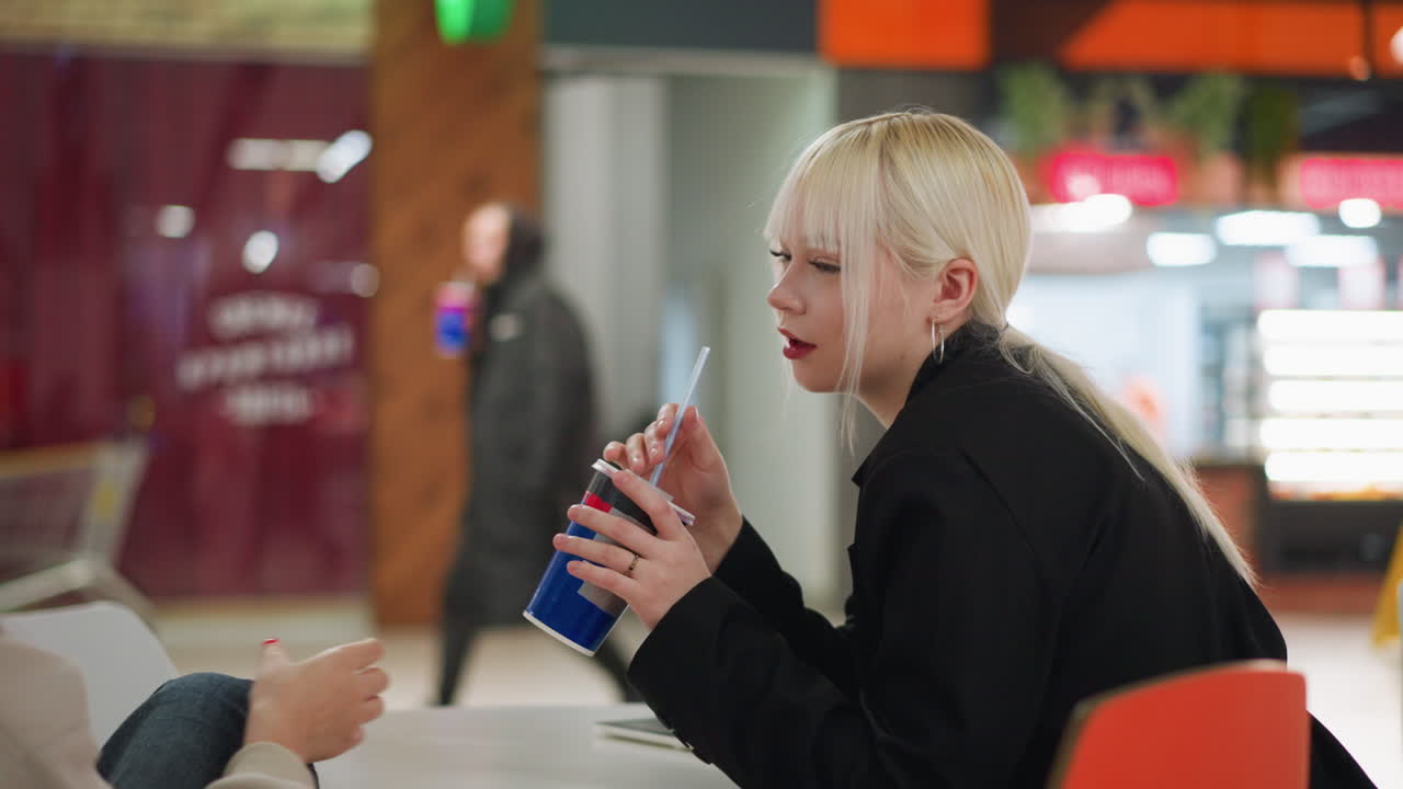 Two young ladies sit across table inside modern cafe, one sipping from drink cup before offering it to her friend while they interact and laugh, blurred passerby in background