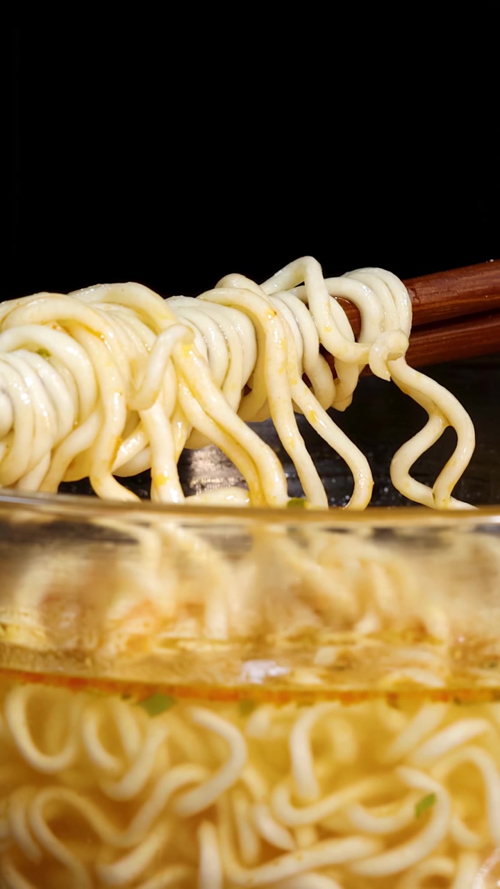 黒い背景の中、手がガラスのスープボウルから湯気の立つインスタントラーメンを木製の箸で持ち上げている。