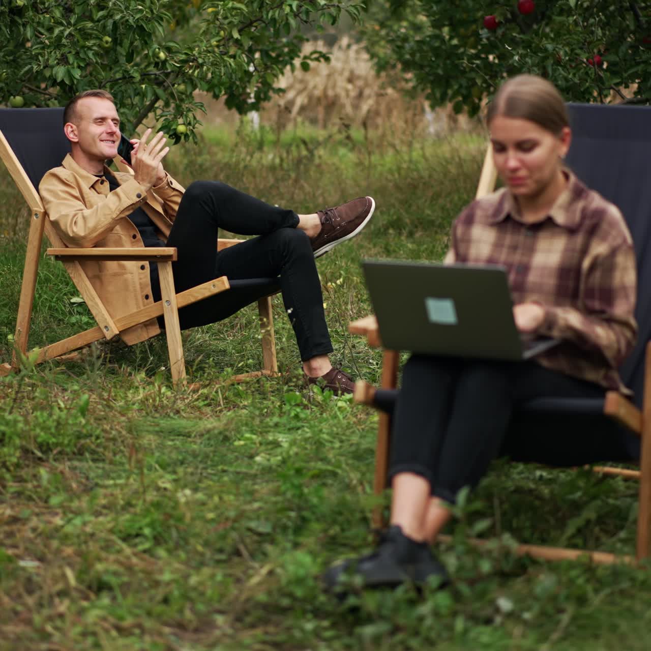 Married couple working online in their garden. Woman works on laptop and man speaking on the phone. Family freelancers working remotely