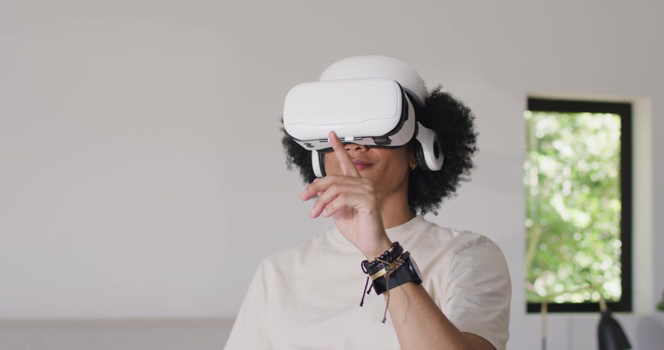 Uomo afroamericano tocca il visore VR e poi gesticola per manipolare oggetti VR a casa vicino alla finestra.