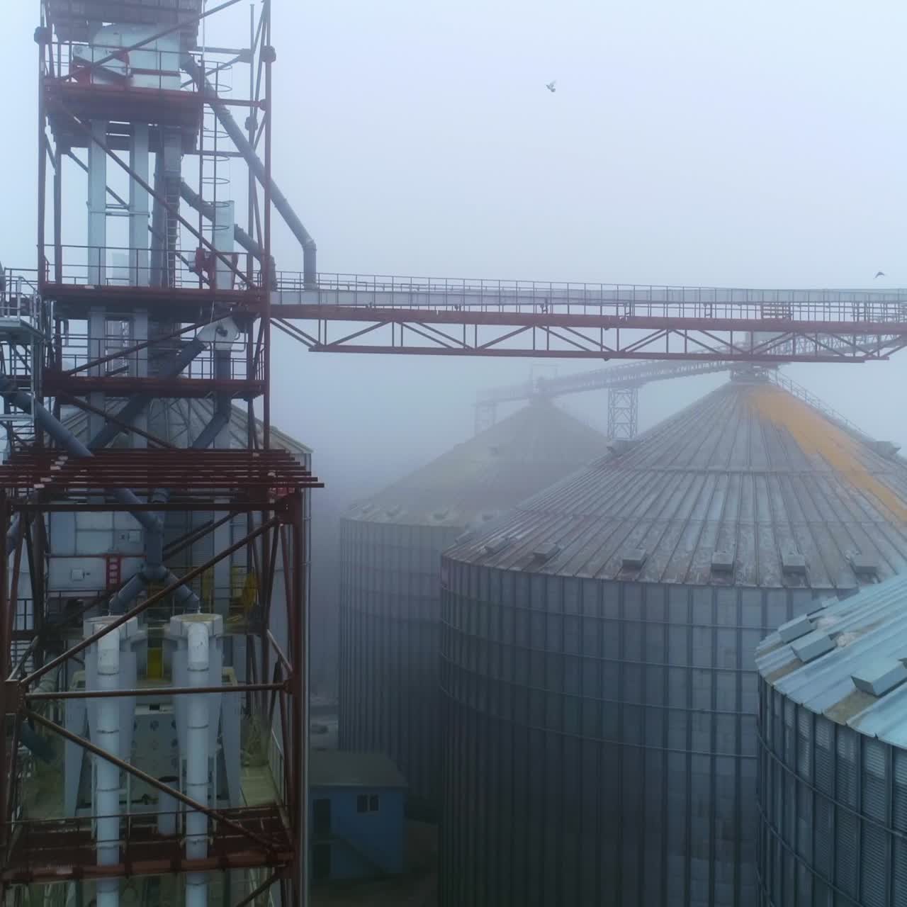 Exterior of powerful agricultural plant on winter foggy day. Metal supports connecting the tanks of elevator. Drone footage