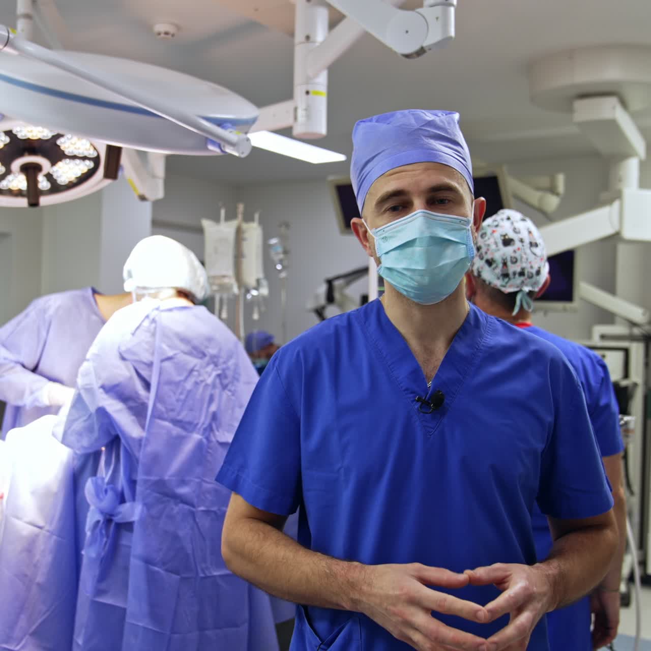 Modern technically equipped surgery room with a large team of professional working in. Doctor in mask speaks to camera. Group of medics at operational table