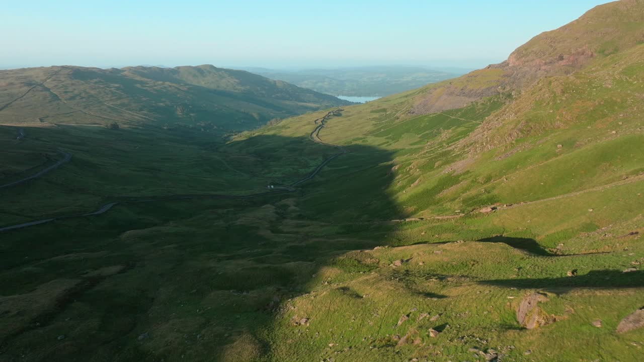 Flying close to mountains illuminated by early morning sunshine. With mountain pass road snaking towards distant lake Windermere. Summer. Kirkstone Pass, Lake District, Cumbria, UK