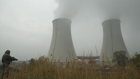 Man-in-a-protective-suit-walks-through-the-meadow-in-front-of-the-power-plant-with-a-dosimeter