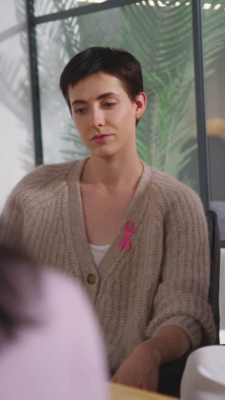 Vertical Video Of Young Woman Wearing Pink Breast Cancer Awareness Ribbon Meeting And Talking At Therapy Support Group For Cancer Treatment Patients With Counsellor 3