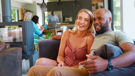 Pareja-Multirracial-En-El-Sofá-De-Casa-Juntos-Viendo-Televisión-Con-Una-Familia-Multigeneracional-En-Segundo-Plano