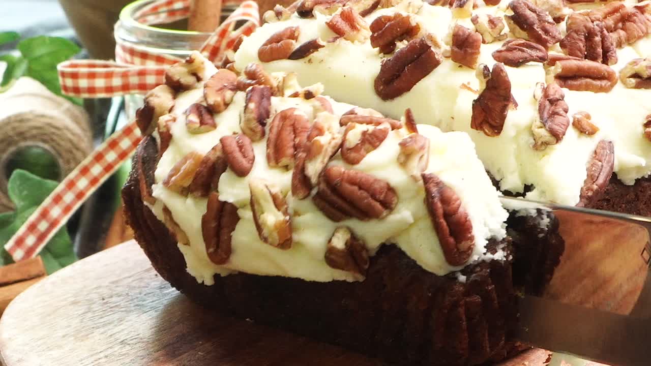 Baking a carrot cake loaf with cream cheese icing and topped with roasted salted pecans. Making a Autumn dessert with carrots, flour, sugar, nuts and frosting. Slicing a piece of carrot cake bread.
