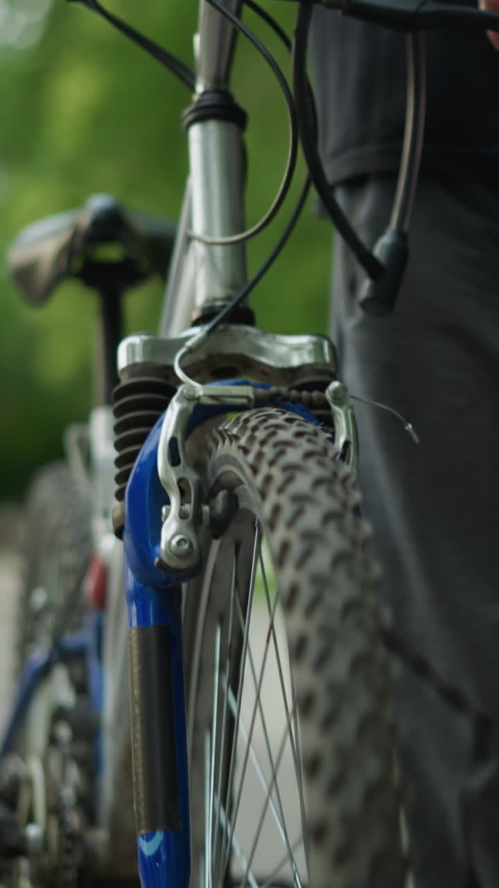 vista parcial de alguien en la parte superior negra y pantalones grises caminando de cerca con bicicleta a lo largo de un camino tranquilo, con vista borrosa de la vegetación y la dama sentada cerca en la distancia