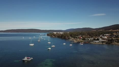 Aerial-drone-pan-around-collection-of-boat-and-yachts-in-deep-blue-sea-surrounded-by-small-mountain-range-and-town-on-clear-sunny-day