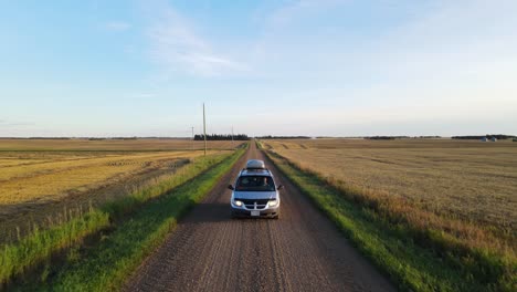 backwards moving aerial tracking shot of a silver mini van driving along a dusty dirt road through the countryside of central alberta