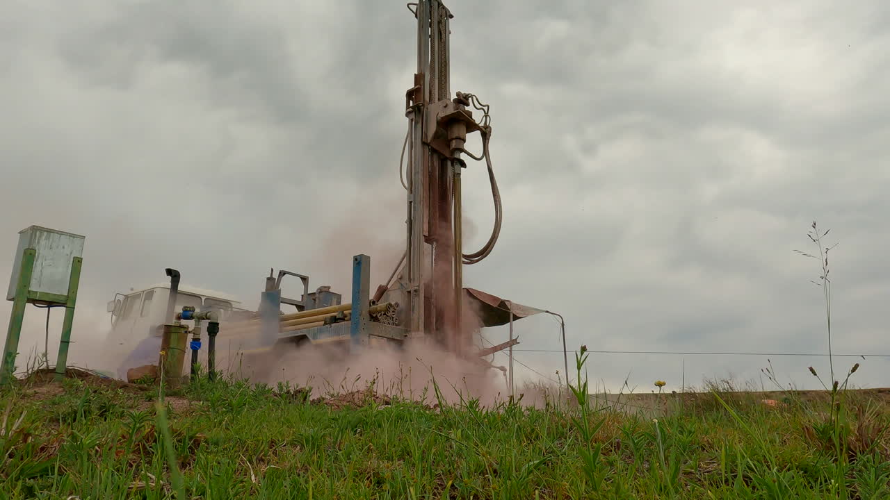 A large drilling machine bores into the ground, kicking up dust under a cloudy sky in a rural field