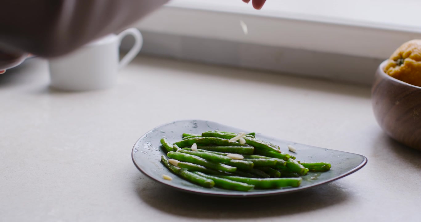 Indian woman pinching garlic and sprinkling over green beans on plate at counter finishing dish
