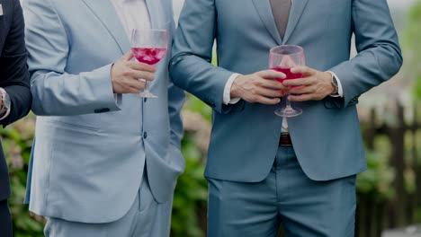 three men in suits holding pink drinks in stemmed glasses at a formal event.