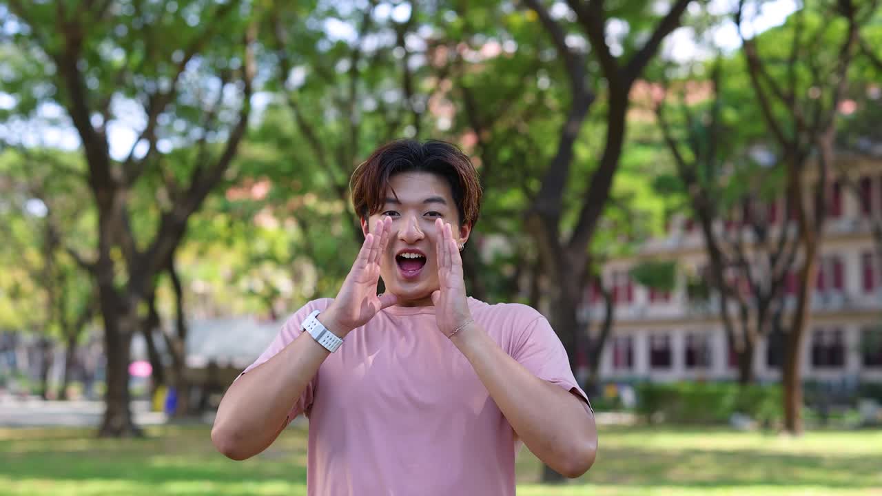 ชายหนุ่มชาวไทยสวมเสื้อเชิ้ตสีชมพูกำลังตะโกนเสียงดังพร้อมทำมือป้องปากกลางแจ้ง