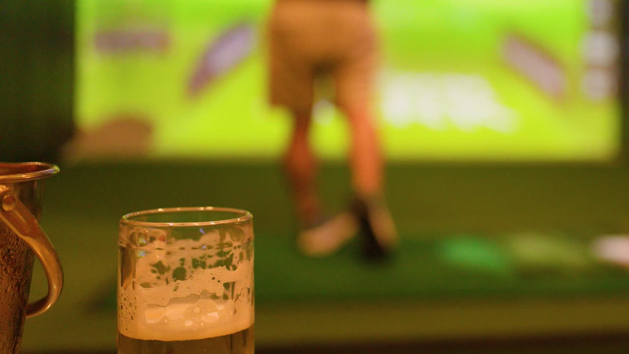 Un uomo esegue uno swing con una mazza da golf in un simulatore indoor dietro un bicchiere di birra fredda