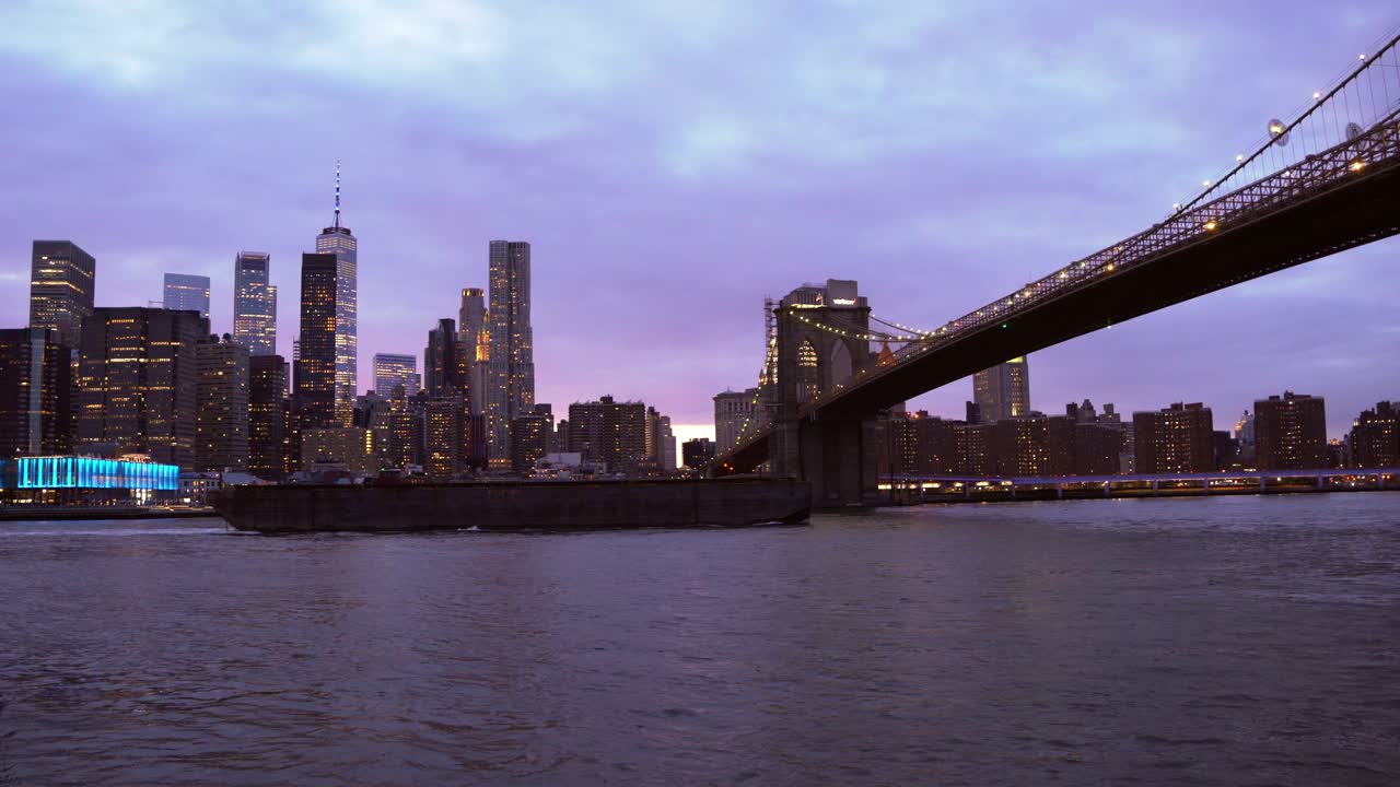 Metraje único de una barcaza pasando bajo el Puente de Brooklyn al atardecer con Nueva York de fondo.