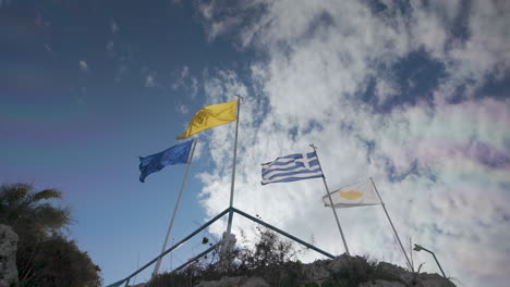 flags of cyprus, greece, and another unidentifiable flag flying high on a pole atop a rocky outcrop against a backdrop of blue sky and clouds