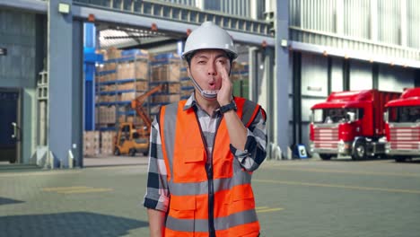 asian male engineer with safety helmet yelling with hand over mouth while standing , outside of logistics distributions warehouse