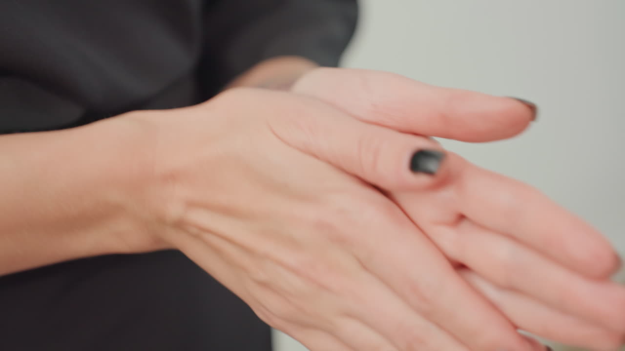 Donna caucasica che si spalma la crema sulle mani. La sequenza ravvicinata mostra i palmi, le dita e le unghie smaltate di nero mentre massaggia la crema, ammorbidisce le cuticole e dimostra la cura delle mani in un luminoso studio al chiuso.