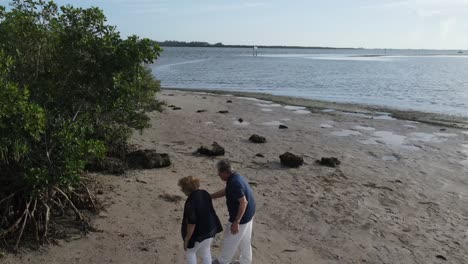 aerial-of-senior-citizen-couple-walking-the-shoreline-of-the-ocean,-exploring-the-beach