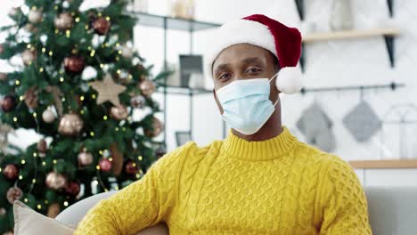 Handsome-Joyful-Man-In-Mask-And-Santa-Hat-Sitting-At-Home-Near-Decorated-With-Lights-Christmas-Tree,-Looking-At-Camera-And-Smiling