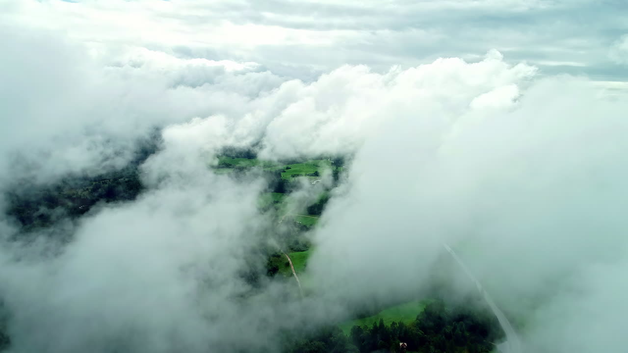 Vivid lush green meadows neighbouring with forests growing on both sides of the road are seen behind the clouds while flying above