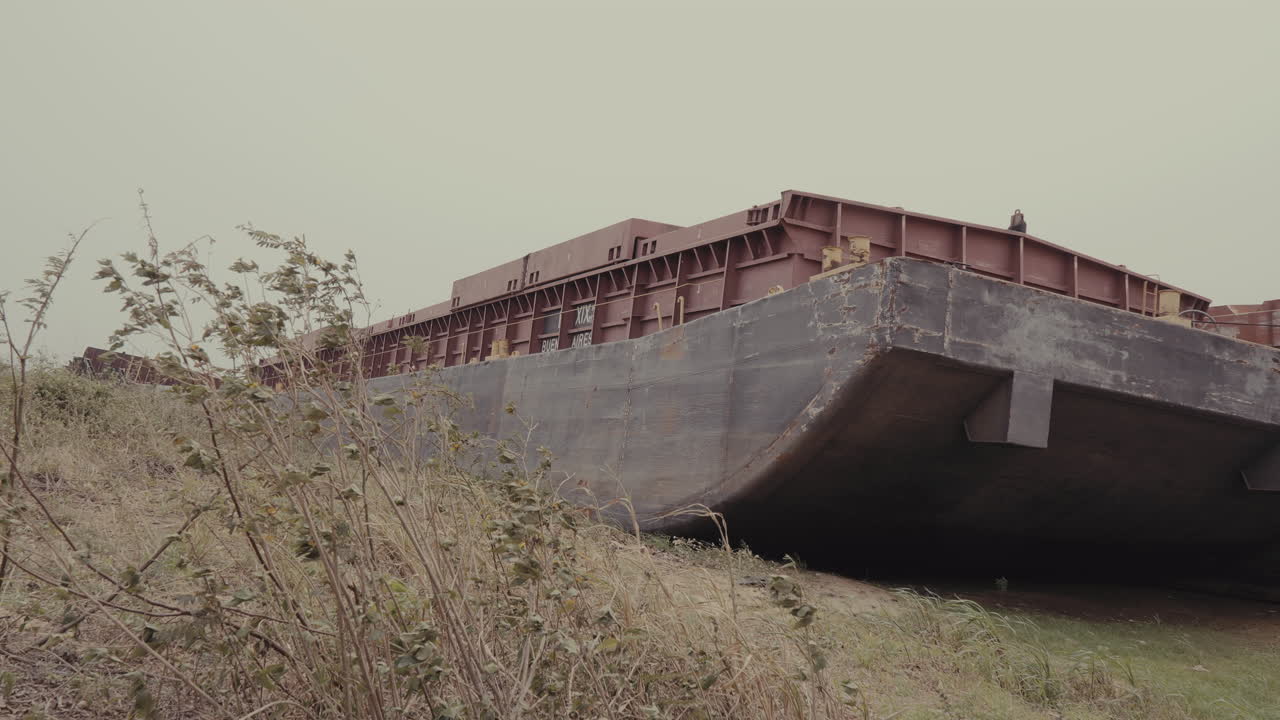 Abandoned Barge on Overgrown Riverbank: Industrial Decay and Nature’s Reclamation. The barge's worn, faded paint indicate years of exposure and neglect