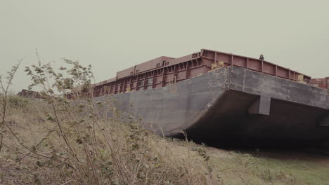 abandoned barge on overgrown riverbank: industrial decay and nature’s reclamation. the barge's worn, faded paint indicate years of exposure and neglect